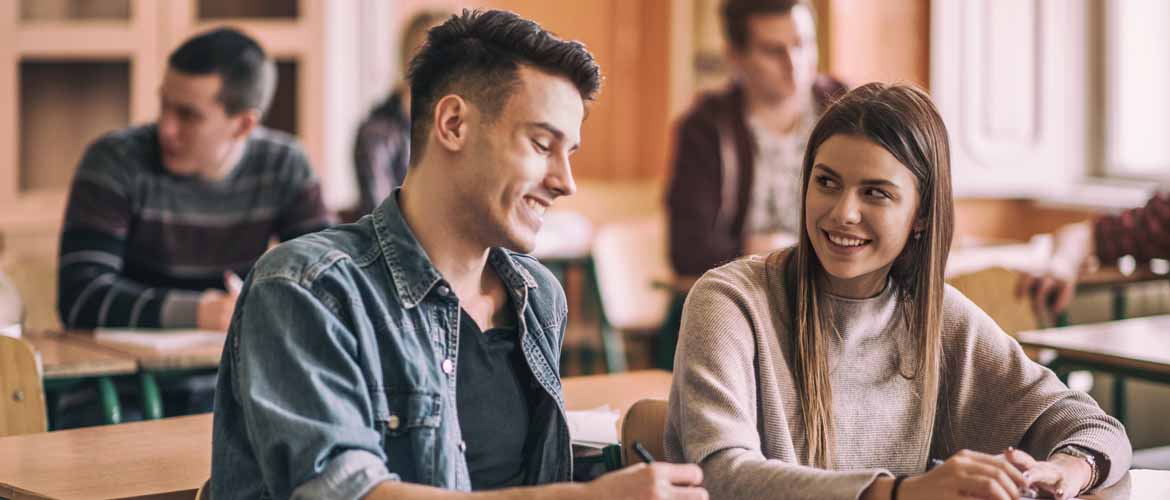 a young man and a young woman sitting next to each other in a classroom with other students in the background