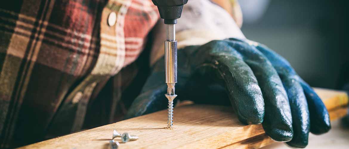 close-up of gloved hands using a power tool to drill a screw into a piece of wood