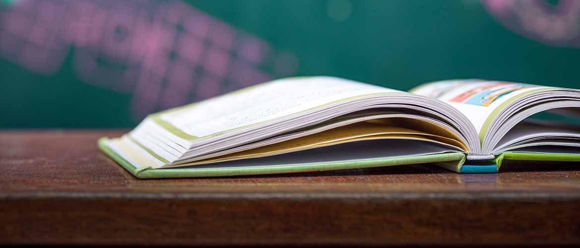 an open book sitting on a school desk in a classroom with a blackboard in the background