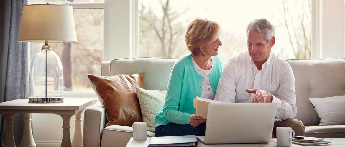 an older couple referencing a computer screen and folder while having a discussion in their living room