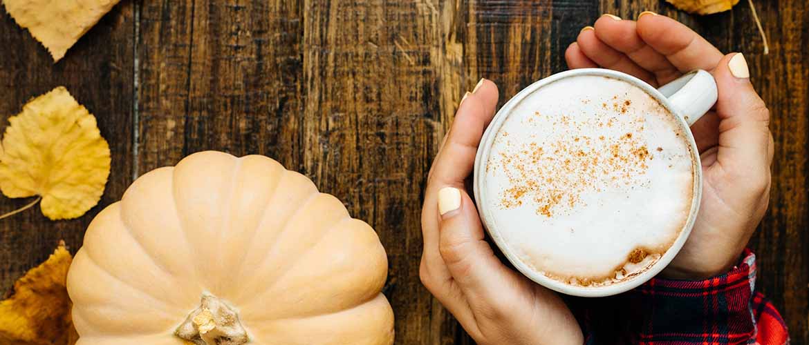 a person holding a latte on a wooden table with a pumpkin and leaves sitting on it